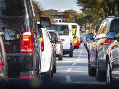 Uitzicht vanuit een auto met een file op een zonnige dag, met focus op de achterlichten van nabijgelegen voertuigen en de Mobiliteitsalliantie-borden.