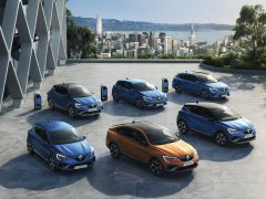 Een reeks van zes elektrische auto's van Renault E-Tech, in verschillende modellen en kleuren, overdag tentoongesteld op een terras met uitzicht op de skyline van de stad aan de baai.