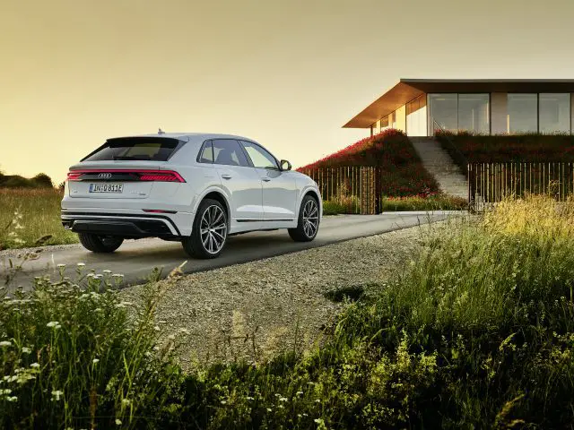 Een witte Audi Q8 TFSI e quattro geparkeerd op een onverharde weg nabij een modern huis met grote ramen bij zonsondergang.