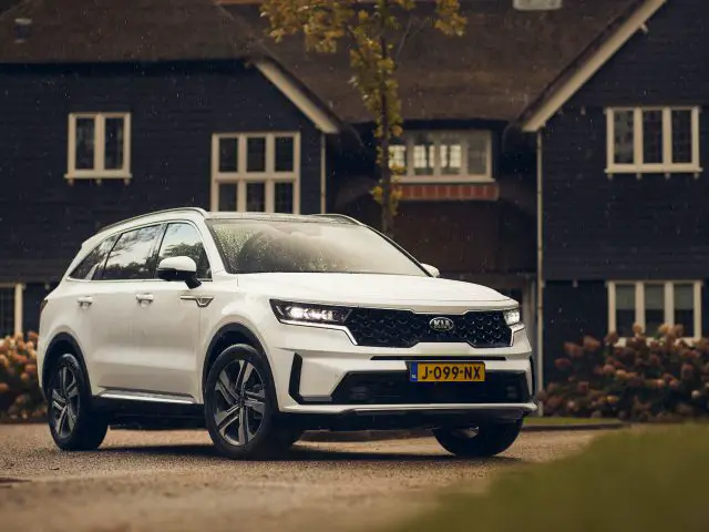 Een witte Kia Sorento hybride SUV geparkeerd voor een traditioneel huis met een rieten dak, op een natte oprit omringd door herfstbladeren.