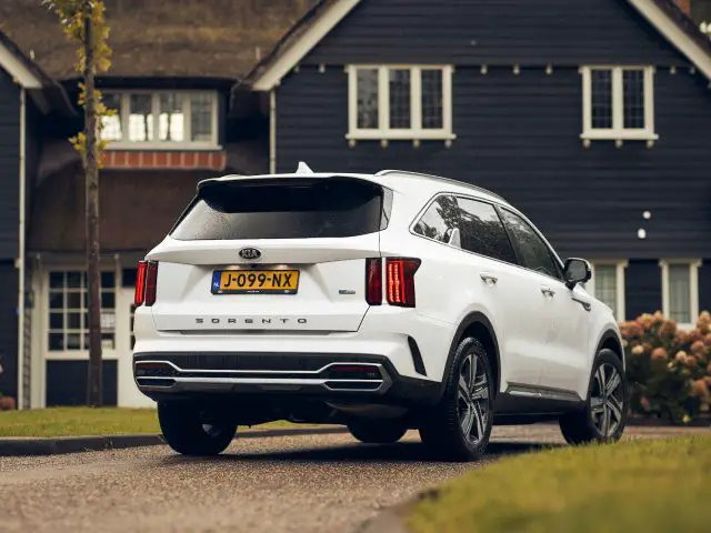 Een witte Kia Sorento Hybrid geparkeerd op een verharde oprit voor een huis met een rieten dak, met de achterklep open.