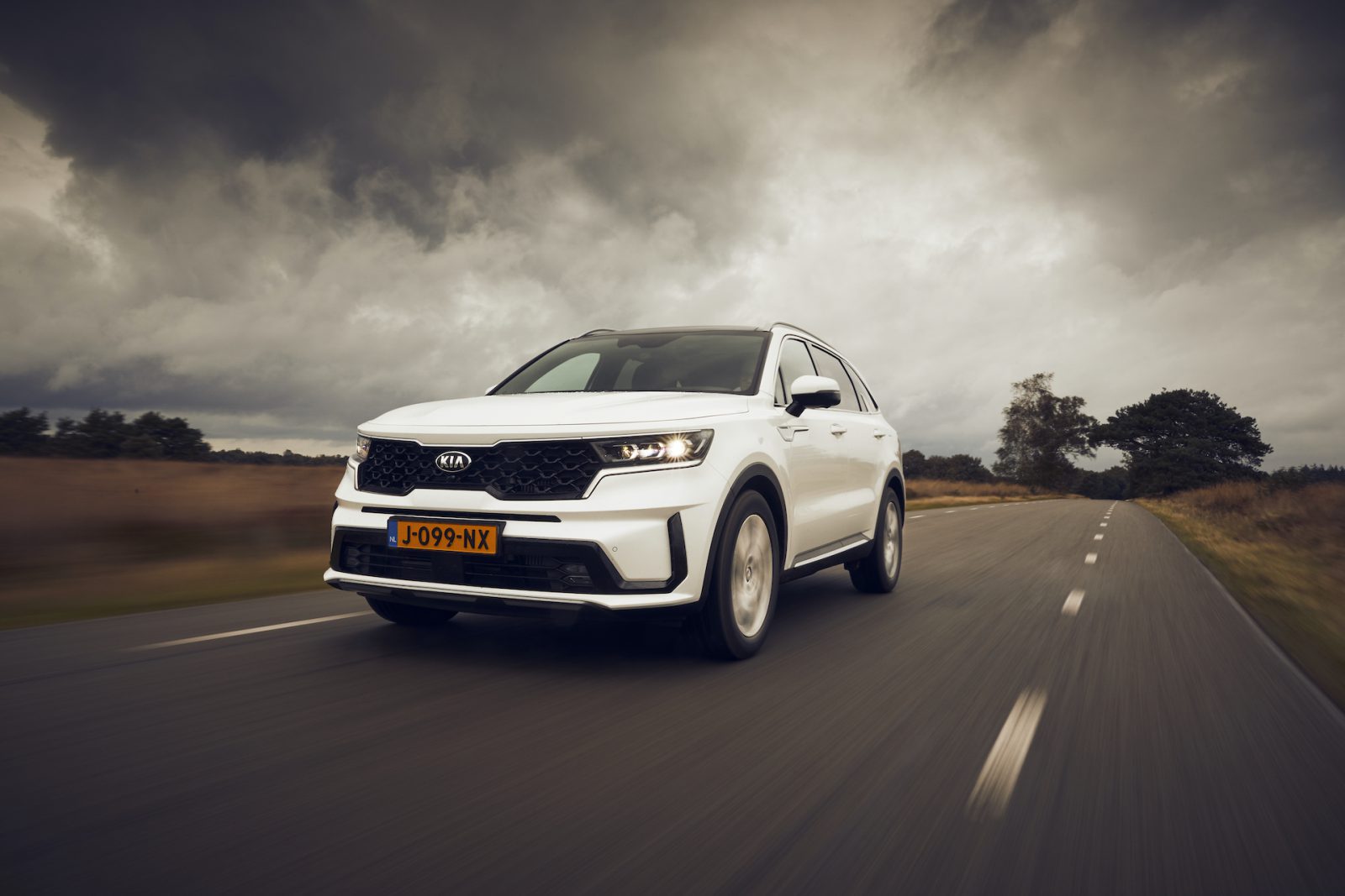 Een witte Kia Sorento Hybrid SUV rijdt op een weg met een bewolkte lucht op de achtergrond.