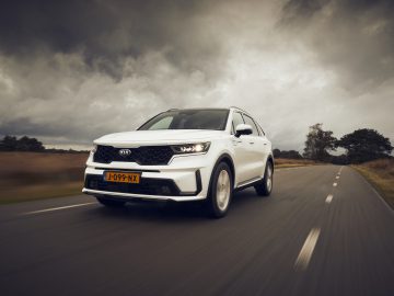 Een witte Kia Sorento Hybrid SUV rijdt op een weg met een bewolkte lucht op de achtergrond.