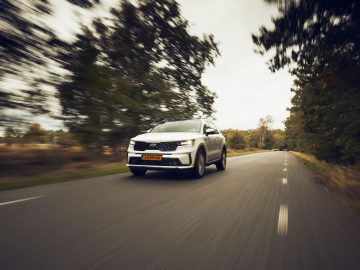 Een witte Kia Sorento Hybrid die snel rijdt op een asfaltweg omringd door herfstbomen, wat een bewegingsonscherpte-effect demonstreert.