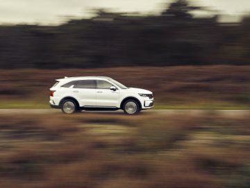 Een witte Kia Sorento Hybrid SUV in beweging, vastgelegd met bewegingsonscherpte op een landelijke weg omgeven door gras en bomen.