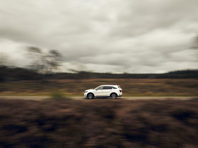 Een witte Kia Sorento Hybrid rijdt over een landelijke weg met vage bomen op de achtergrond, wat de beweging benadrukt.