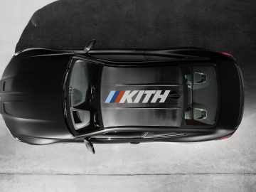 Luchtfoto van een zwarte BMW M4 Competition x KITH-auto met een opvallend kith-logo op het dak, geparkeerd op een grijs betonnen oppervlak.