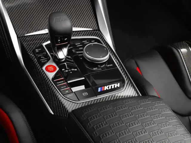 Close-up van de middenconsole van een BMW M4 Competition x KITH met een versnellingspook, verschillende bedieningsknoppen en een draaiknop, allemaal tegen een achtergrond van koolstofvezel.