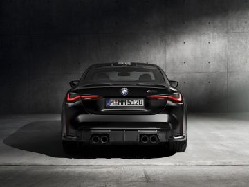 Achteraanzicht van een zwarte BMW M4 Competition x KITH-auto geparkeerd tegen een betonnen muur, met de nadruk op de dubbele uitlaten en het M4-embleem.