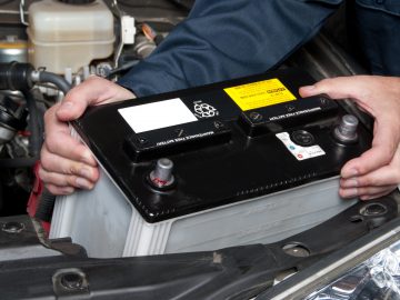 Een persoon die een spijkerjasje draagt, installeert een zwarte Accu-autobatterij onder de motorkap van een voertuig.