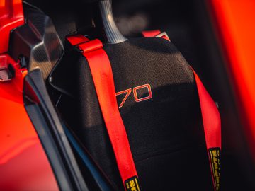Close-up van een zwarte Donkervoort D8 GTO-JD70 R autostoeltje met rode veiligheidsgordels, met een in wit geborduurd '70'-logo op de hoofdsteun, in een