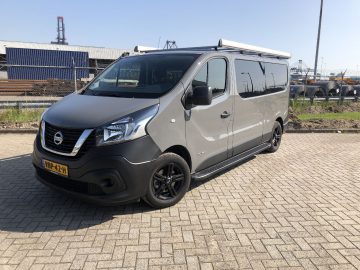 Een Nissan NV300-busje geparkeerd op een geplaveid terrein nabij industriële gebouwen onder een helderblauwe lucht.