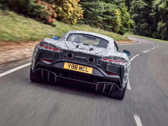 Een zilveren McLaren-sportwagen met prototype-camouflage rijdt op een bochtige weg omgeven door weelderig groen.