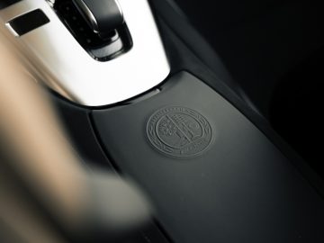 Close-up van een logo in reliëf op de middenconsole van een Mercedes-AMG GT C Roadster, gepositioneerd tussen de bestuurdersstoel en de versnellingspook.