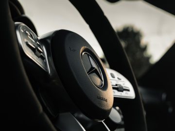 Close-up van het stuur en het dashboard van een Mercedes-AMG GT C Roadster, met de nadruk op het logo, met een wazig auto-interieur op de achtergrond.