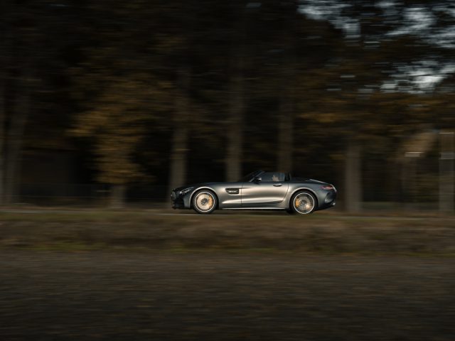 Een zilveren Mercedes-AMG GT C Roadster snelt langs een wazige bosachtergrond en creëert een gevoel van beweging.