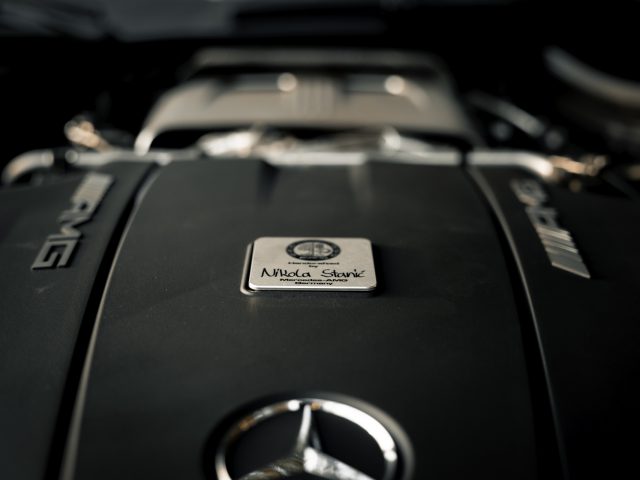 Close-up van een Mercedes-AMG GT C Roadster-motor met het logo en het embleem "AMG Affalterbach" op een gepolijste cover, verlicht door zachte verlichting.
