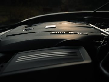 Close-up van een Mercedes-AMG GT C Roadster-motorkap met gedetailleerd logo, verlicht door natuurlijk licht.