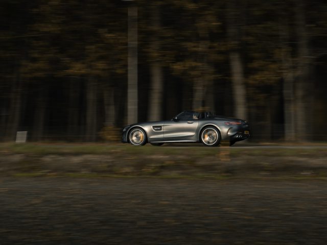 Een zilveren Mercedes-AMG GT C Roadster die met bewegingsonscherpte over een bosweg snelt, wat de hoge snelheid benadrukt.