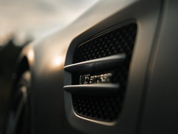 Close-up van de ventilatieopening aan de zijkant van een Mercedes-AMG GT C Roadster met een 'v8 biturbo'-badge, met de nadruk op de getextureerde grille tegen een onscherpe natuurlijke achtergrond.