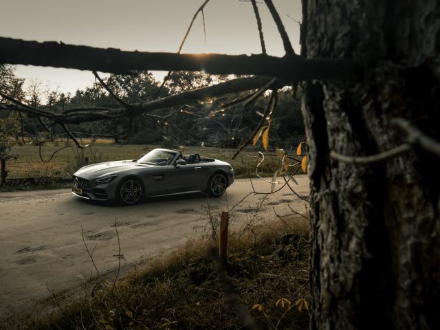 Een zilveren Mercedes-AMG GT C Roadster geparkeerd op een onverharde weg nabij een bosrijke omgeving, gezien door takken op de voorgrond.