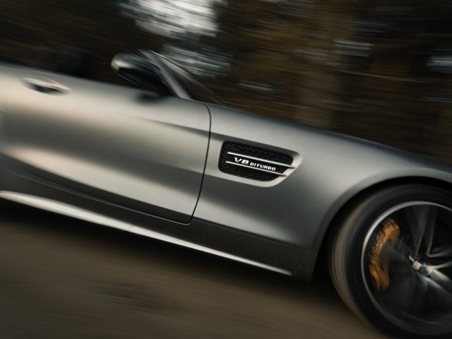 Een zilveren Mercedes-AMG GT C Roadster in beweging, met een "v8 biturbo" -badge op de zijkant, met een onscherpe achtergrond die hoge snelheid aangeeft.
