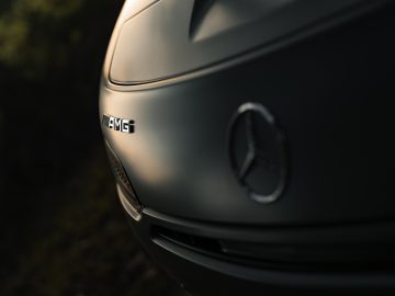 Close-up van de voorkant van een zilveren Mercedes-AMG GT C Roadster, met de nadruk op het logo en het AMG-embleem op een softfocusachtergrond.