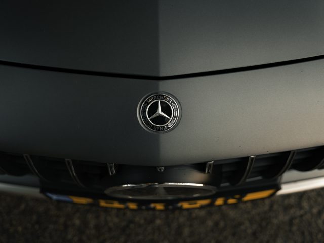 Close-up van een Mercedes-AMG GT C Roadster-embleem op de grijze grille van een voertuig, met gedetailleerde branding en design.