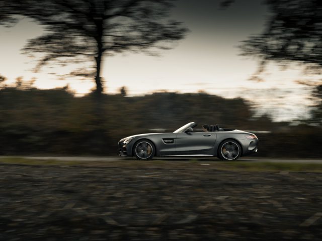 Een zilveren Mercedes-AMG GT C Roadster rijdt in de schemering over een met bomen omzoomde weg, met bewegingsonscherpte en een levendige avondlucht.