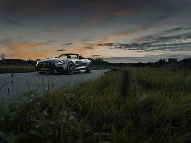 Een Mercedes-AMG GT C Roadster geparkeerd op een verlaten weg in de schemering, met een levendige zonsondergang en bewolkte lucht op de achtergrond.
