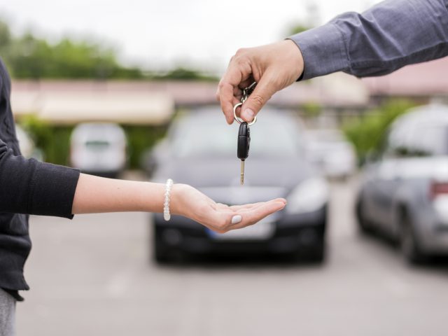 Een persoon overhandigt autosleutels aan de uitgestrekte hand van iemand anders op een parkeerplaats.