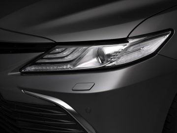 Close-up van de koplamp van een Toyota Camry en een deel van de grille, waarbij het strakke ontwerp en de led-verlichting op een zilverkleurige auto worden benadrukt.