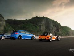Drie auto's geparkeerd op een kustweg: een blauwe sportwagen, een blauwe hatchback en een oranje roadster, met kliffen en een dramatische lucht op de achtergrond, die scènes uit The Grand oproepen