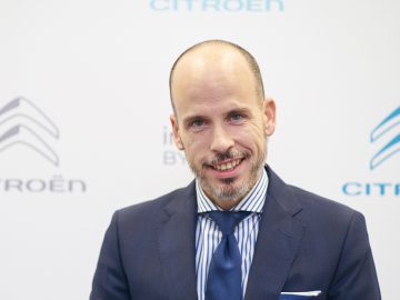 Een man in een blauw pak met een gestreepte stropdas glimlachend tegen een achtergrond met het Citroën-logo.