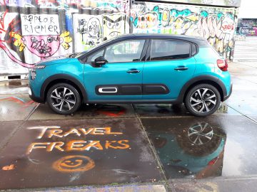 Een blauwgroen Citroën C3 hatchback geparkeerd voor een met graffiti bedekte muur, met 'reisfreaks' geschilderd op de natte grond.