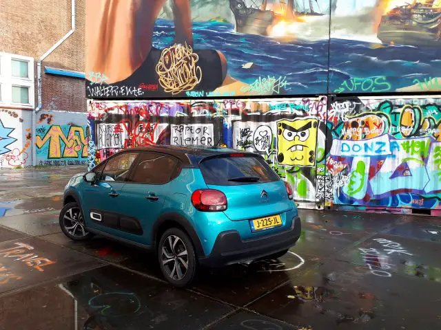 Een Citroën C3 geparkeerd voor een muur bedekt met kleurrijke graffiti onder een natte betonnen vloer.