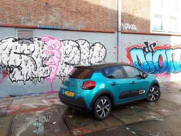 Een blauwgroen Citroën C3 geparkeerd voor een met graffiti bedekte muur op een zonnige dag. De graffiti bevat verschillende kleurrijke tags en ontwerpen.