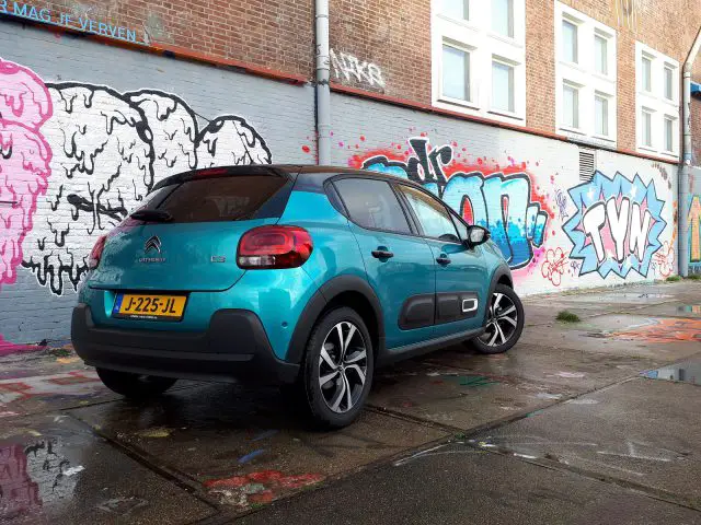 Een blauwgroen Citroën C3 geparkeerd voor een met graffiti bedekte muur in een geplaveide straat.