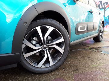 Blauwe Citroën C3 geparkeerd op nat asfalt met een close-up van zijn stijlvolle lichtmetalen velg, vlakbij een muur vol graffiti.