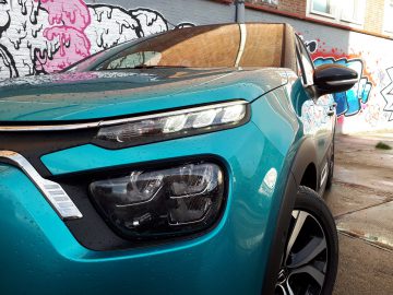Close-up van een blauwgroen Citroën C3 met led-koplampen en glanzende afwerking, geparkeerd voor een met graffiti bedekte muur.