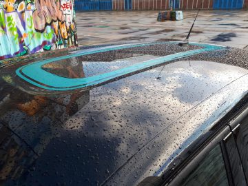 Het natte Citroën C3-dak weerspiegelt een met graffiti bedekte muur, met een opvallende blauwe streep en verspreide regendruppels zichtbaar.