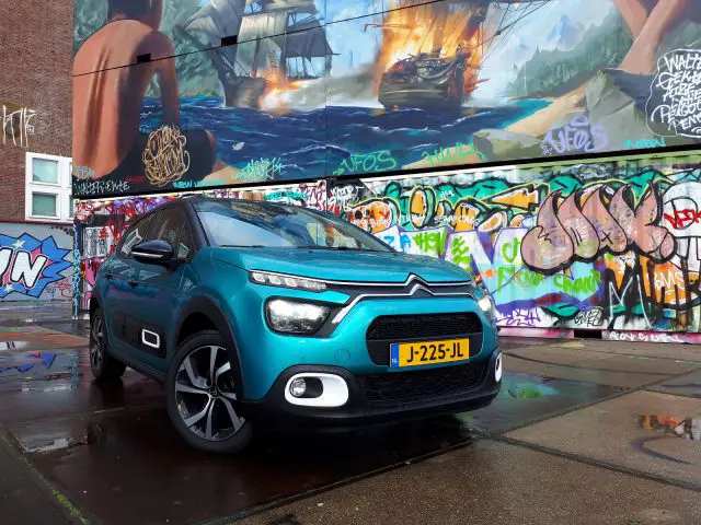 Turquoise Citroën C3 geparkeerd in een natte straat, met kleurrijke graffitikunstwerken op de achtergrond.