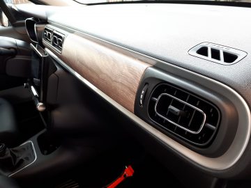 Interieur van een Citroën C3 met het dashboard met ventilatieopening, houten bekleding en een dashboardkastje, verlicht door natuurlijk licht.