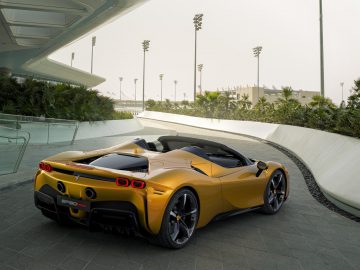 Een gele Ferrari SF90 Spider geparkeerd op een bochtige weg naast een modern gebouw met palmbomen en lichtmasten op de achtergrond.