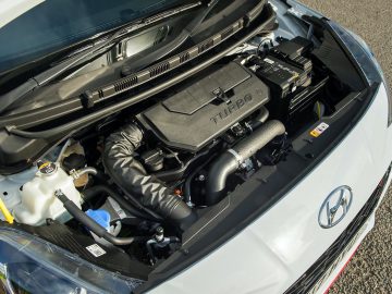 Motorruimte van een witte Hyundai i10 N Line, met een gedetailleerd aanzicht van een turbomotor met zichtbare componenten en labels.