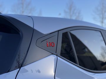 Close-up van een zilveren Hyundai i10 N Line met de zijspiegel en een rode 'i10'-badge op de raamstijl in focus, met een onscherpe achtergrond.