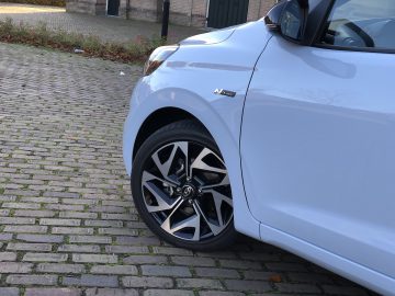 Witte Hyundai i10 N Line geparkeerd in een bakstenen straat, met de nadruk op het voorwiel met een zichtbaar logo op de wieldop.