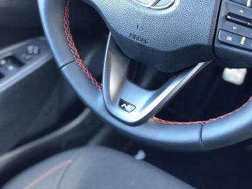 Close-up van een Hyundai i10 N Line-stuur met rood stiksel en een zichtbare spraakopdrachtknop op de linkerspaak.
