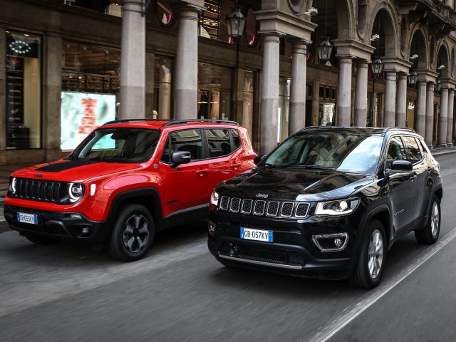 Twee Jeep-SUV's, een rode en een zwarte, rijden zij aan zij door een stadsstraat met klassieke architectuur op de achtergrond.
