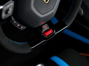 Close-up van het stuur van een Lamborghini Huracán STO met een rood verlichte start/stop-motorknop, geplaatst in een modern dashboard met blauwe accenten.